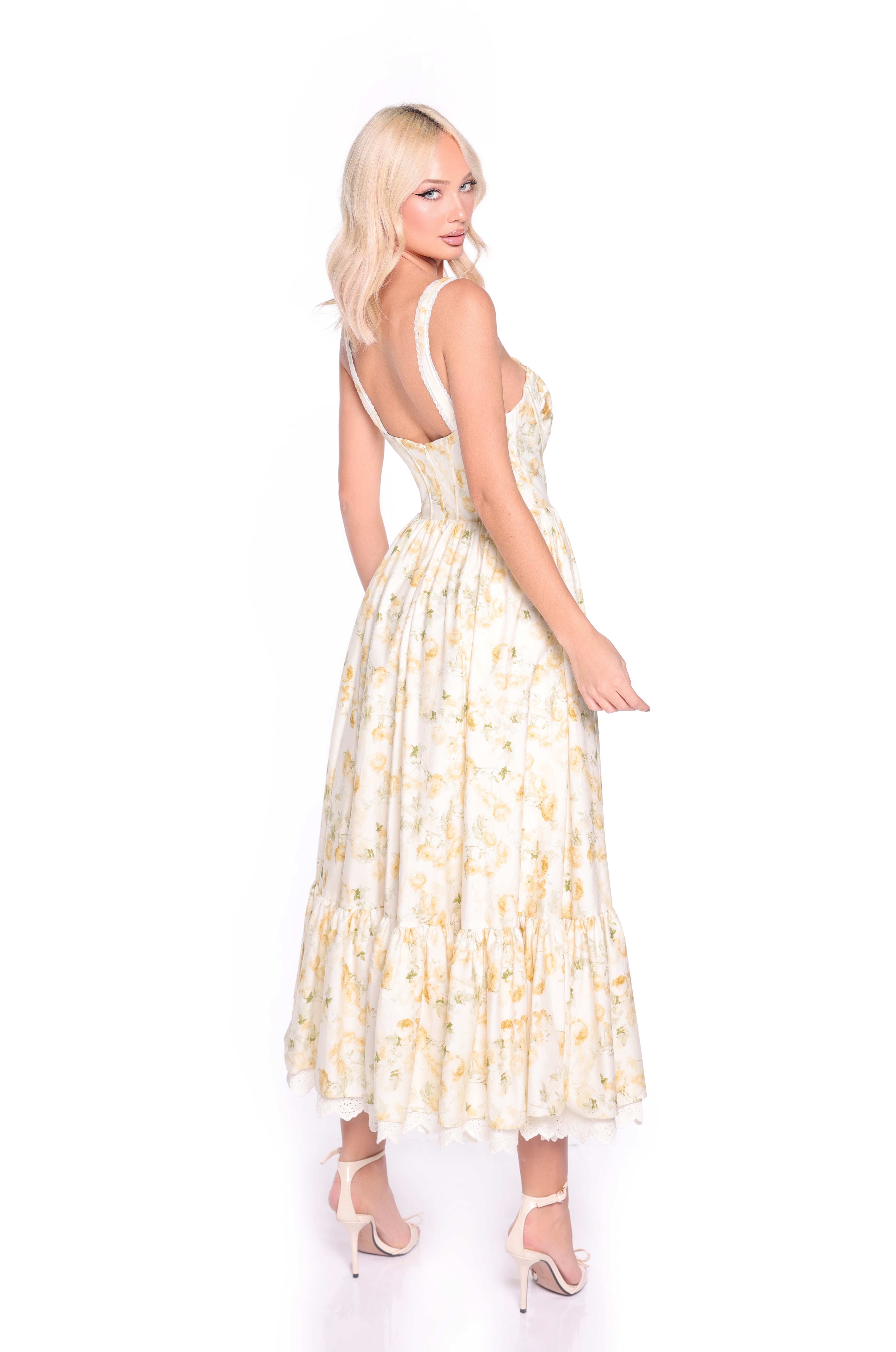 A woman with blonde hair wears the Buttercream Floral Casual Midi Dress: Marigold—a sleeveless, corset-bodice yellow floral midi dress—with white high-heeled sandals, standing against a plain white background.